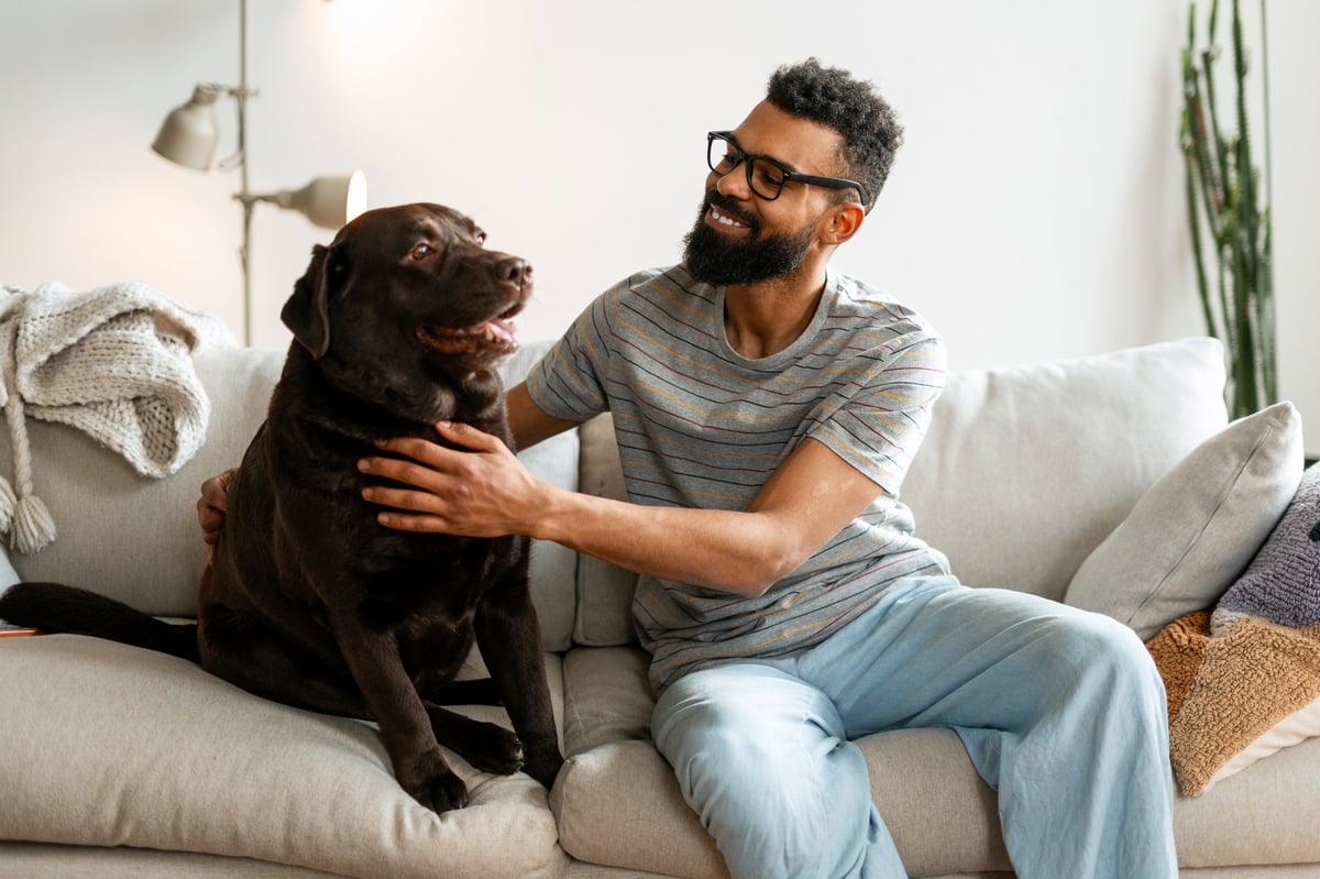 A man sitting on a living room sofa petting his dog, the two of them relaxed together at home